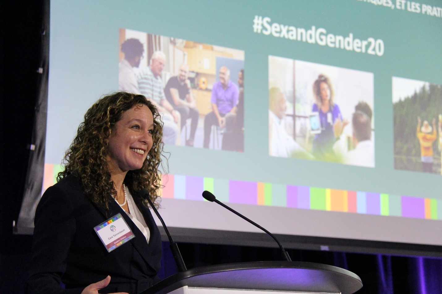 Dr. Cara Tannenbaum, standing at a podium, looking at something out of frame and smiling. Behind her is a screen with colourful images and text reading #SexAndGender20.