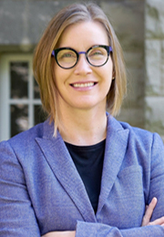 Greta Bauer, PhD, a light-skinned person with shoulder-length, light brown hair with blonde highlights, is outside on a bright day. She is wearing round, black and purple-rimmed glasses and a purple blazer over a black shirt. She is looking towards the camera and smiling, with her arms folded.